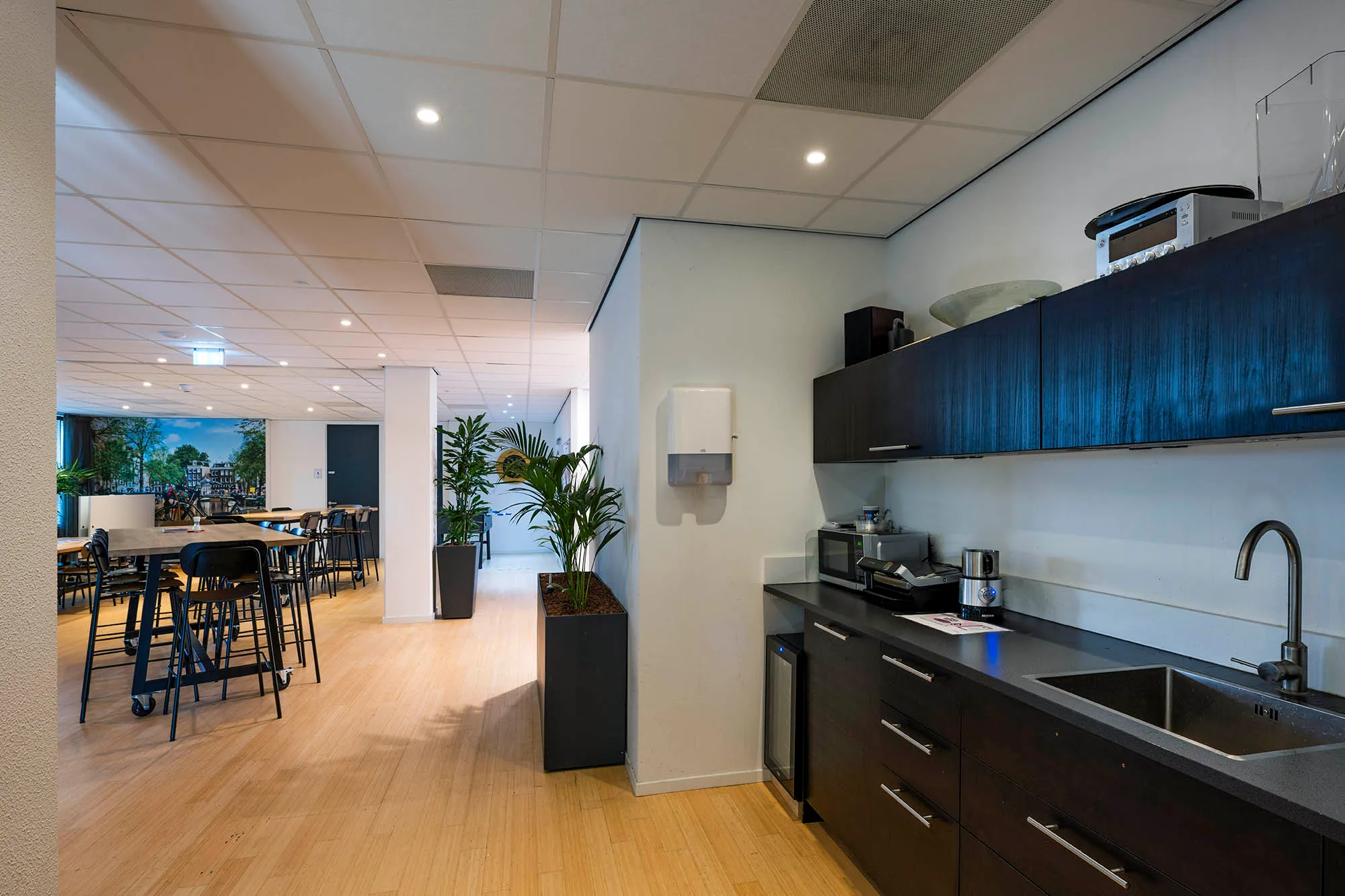 Moderne kantoorruimte met keuken en zithoek met hoge tafels en groene planten.