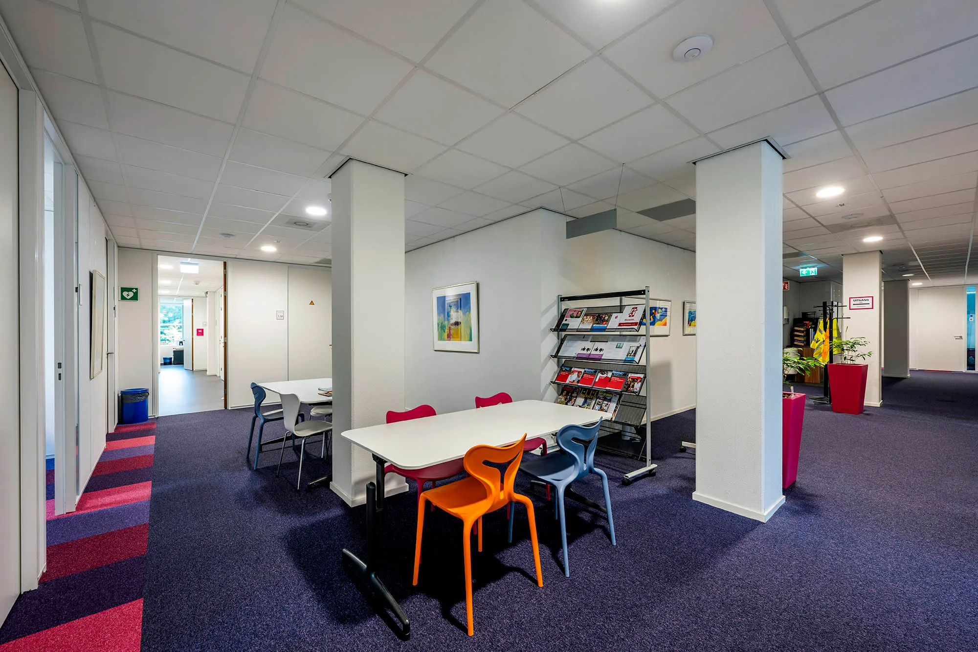Een binnenruimte met een tafel, kleurrijke stoelen en een boekenrek in een kantooromgeving.