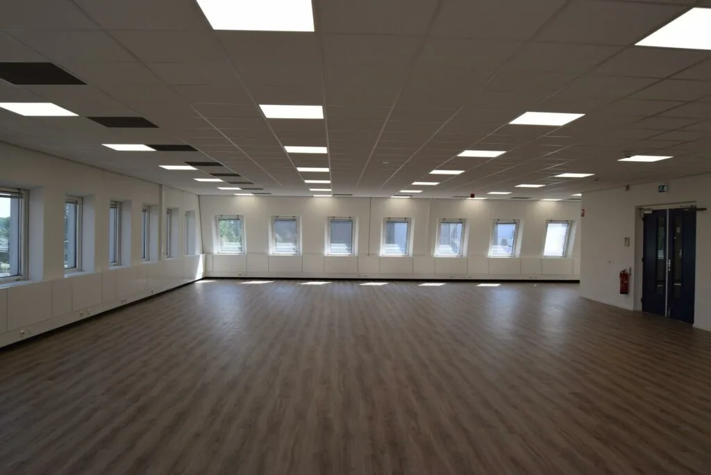 Lichte, lege ruimte met houten vloer, systeemplafond en meerdere ramen in een gebouw aan de Twentestraat.