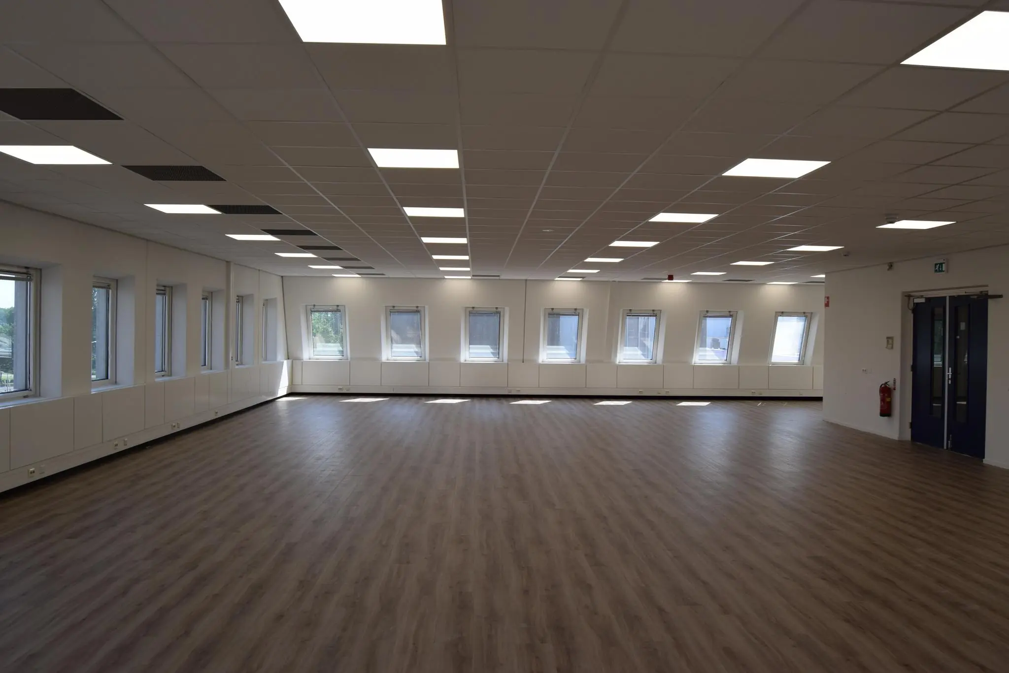 Lichte, lege ruimte met houten vloer, systeemplafond en meerdere ramen in een gebouw aan de Twentestraat.