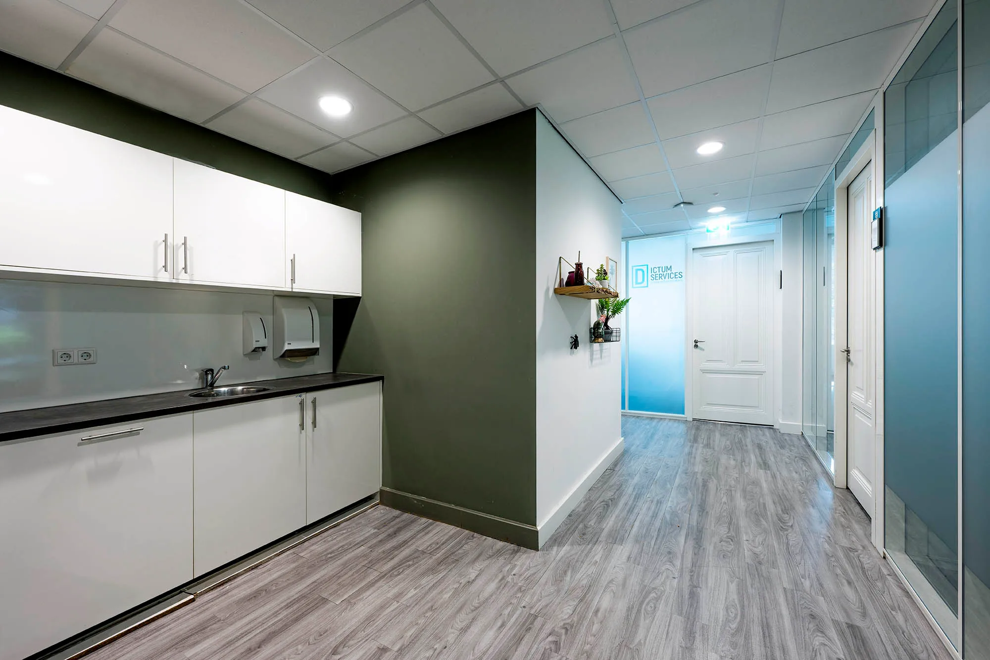 Een moderne kantoorhal met een witte keuken tegen een groene muur en glazen panelen.