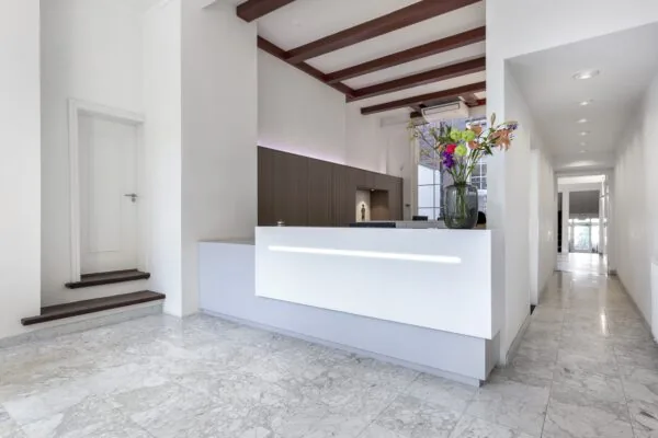 Een moderne receptie met een wit bureau, daarbovenop een vaas met kleurrijke bloemen, in een lichte ruimte met marmeren vloeren.