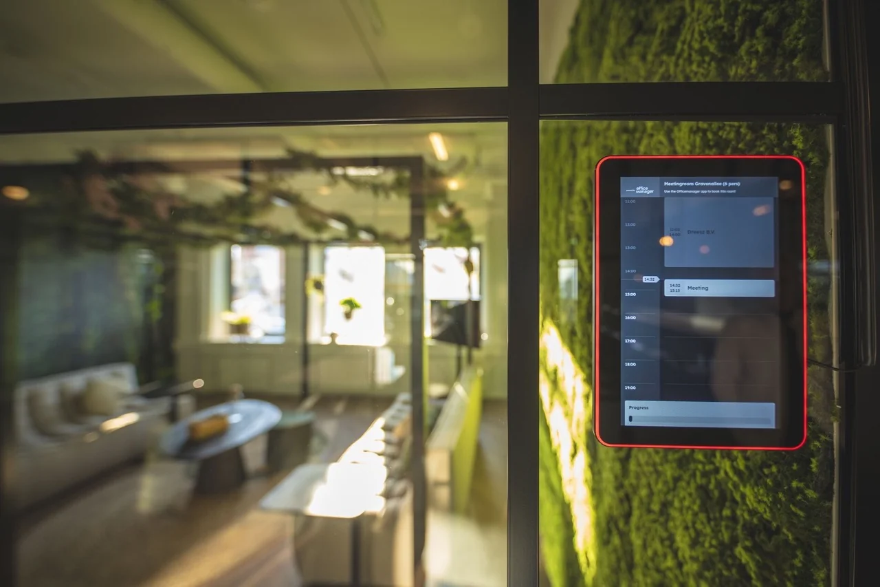 Digitaal reserveringspaneel naast een vergaderruimte met groene moswand en modern interieur.