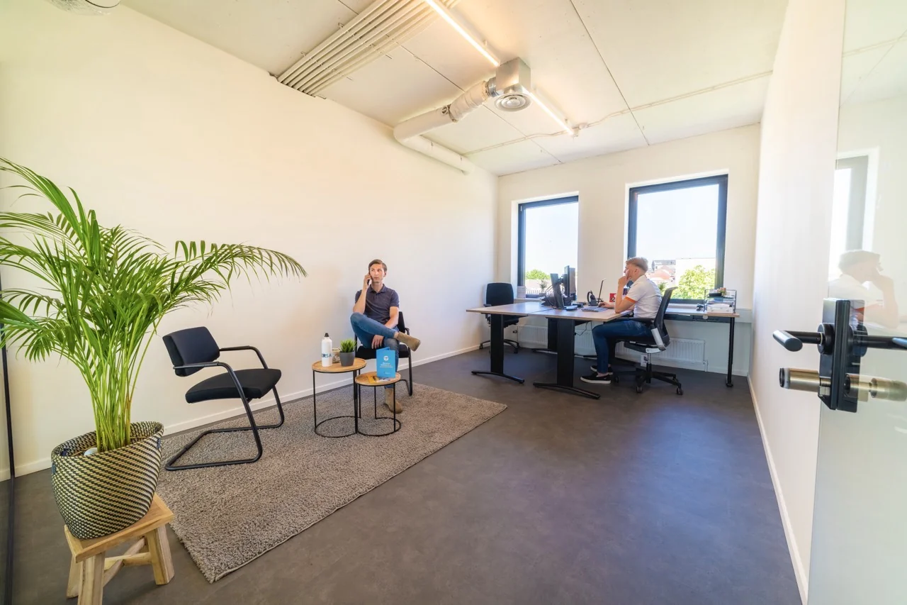 Twee mannen werken in een modern en licht kantoor met grote ramen en een plant in de hoek.