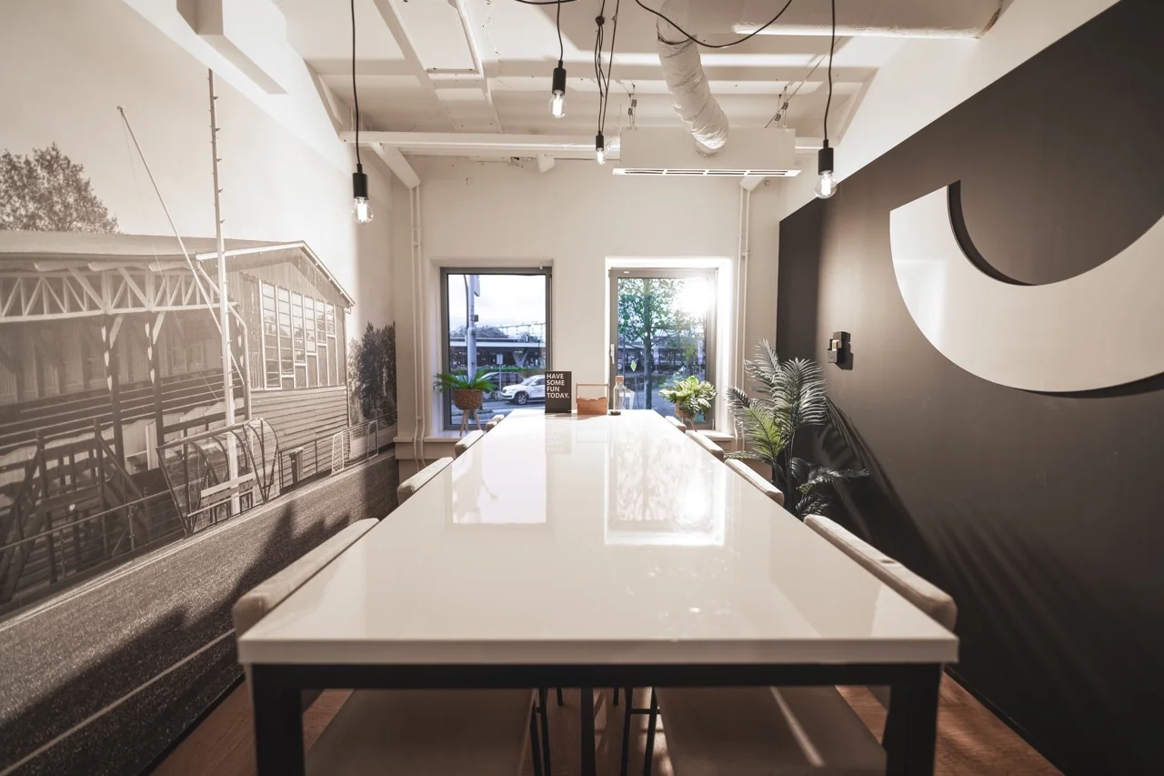 Moderne vergaderruimte met lange witte tafel, muurdecoratie en grote ramen met zicht op buiten.
