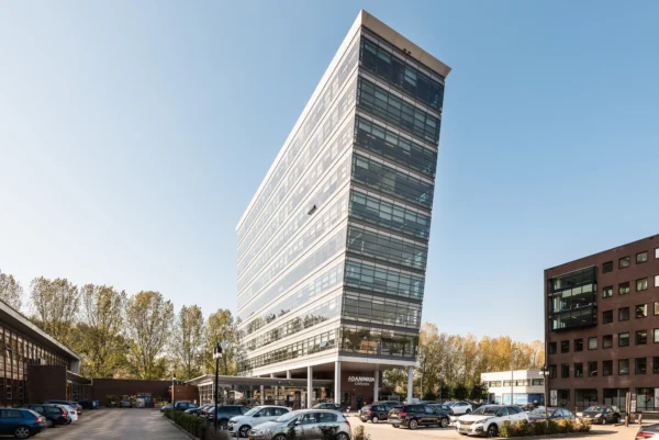 Schuin uitlopende moderne kantoorgebouw met glazen gevels en een parkeerplaats op de voorgrond.