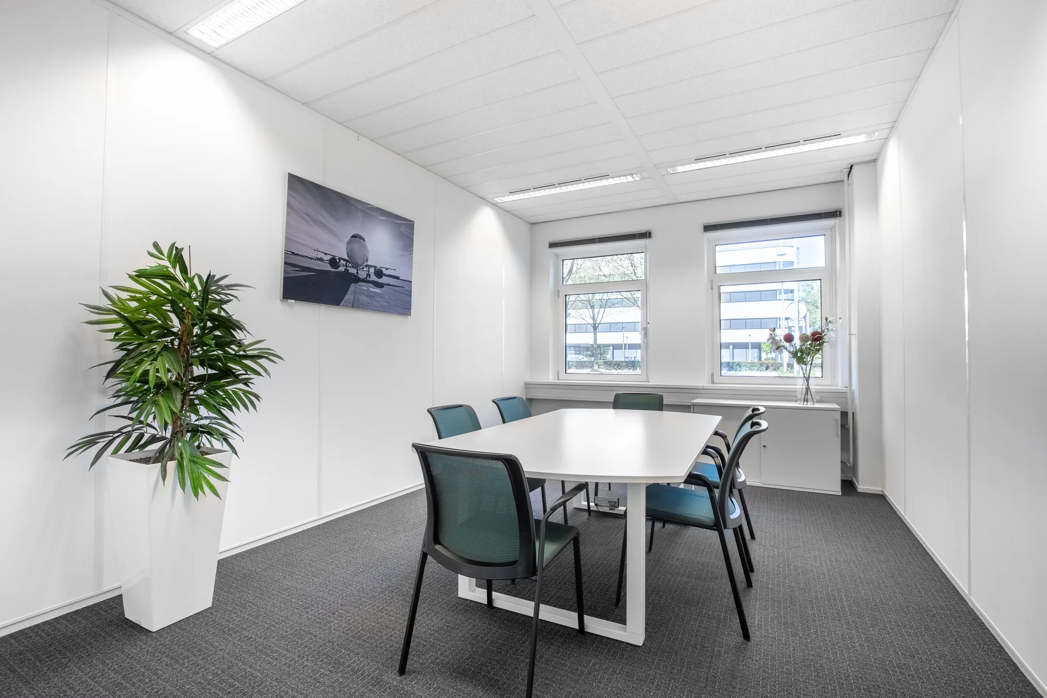 Moderne vergaderruimte met een tafel, zes stoelen, een schilderij van een vliegtuig en grote ramen.