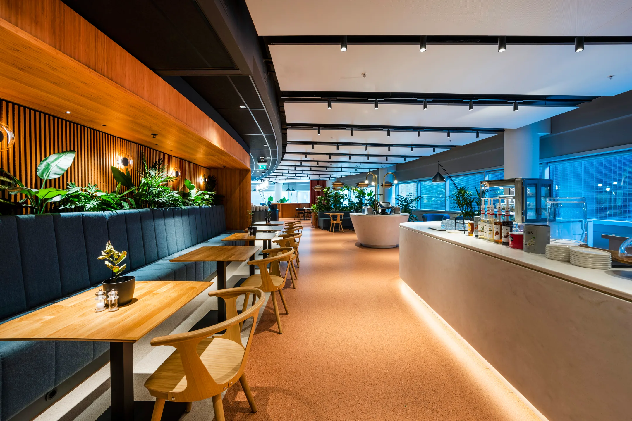 Een modern restaurantinterieur met houten tafels, blauwe zitbanken, planten en een goed verlichte bar.
