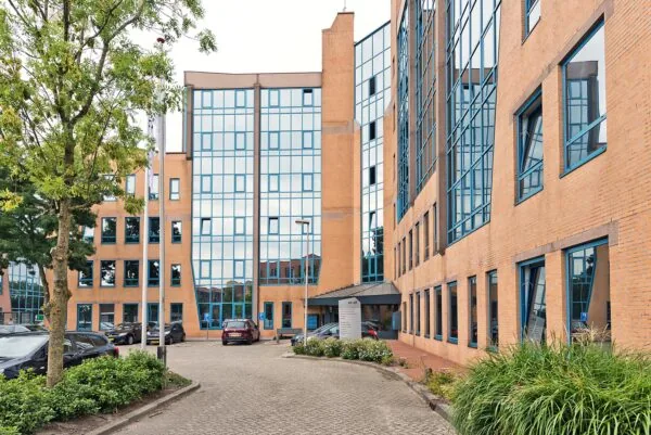 Modern kantoorgebouw aan het Kosterijland met oranje bakstenen, grote blauwe ramen en een parkeerterrein op de voorgrond.