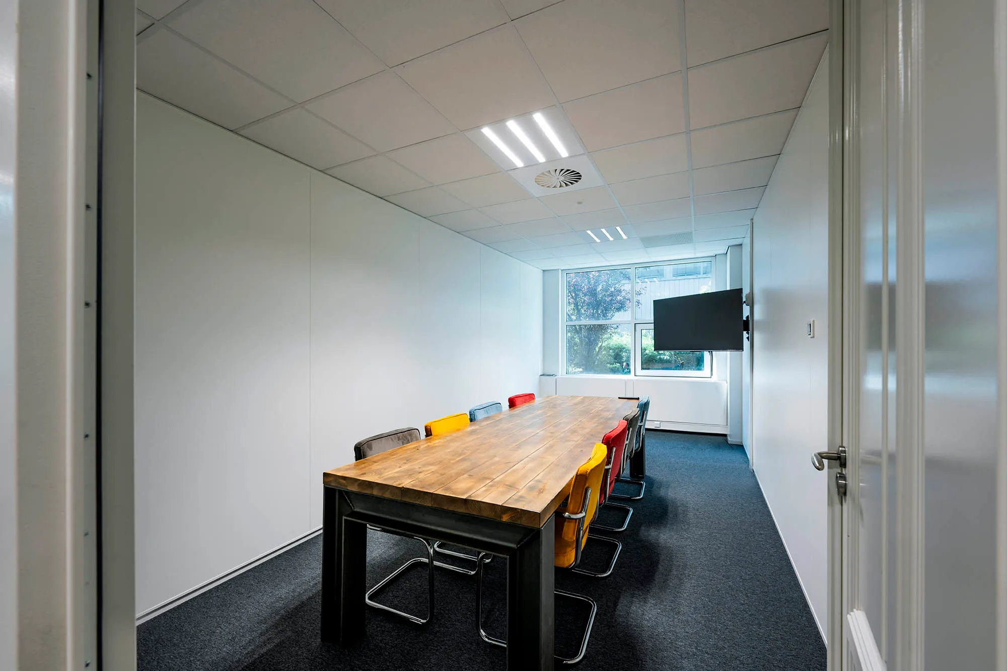 Een vergaderruimte met een grote houten tafel, kleurrijke stoelen en een scherm aan de muur.