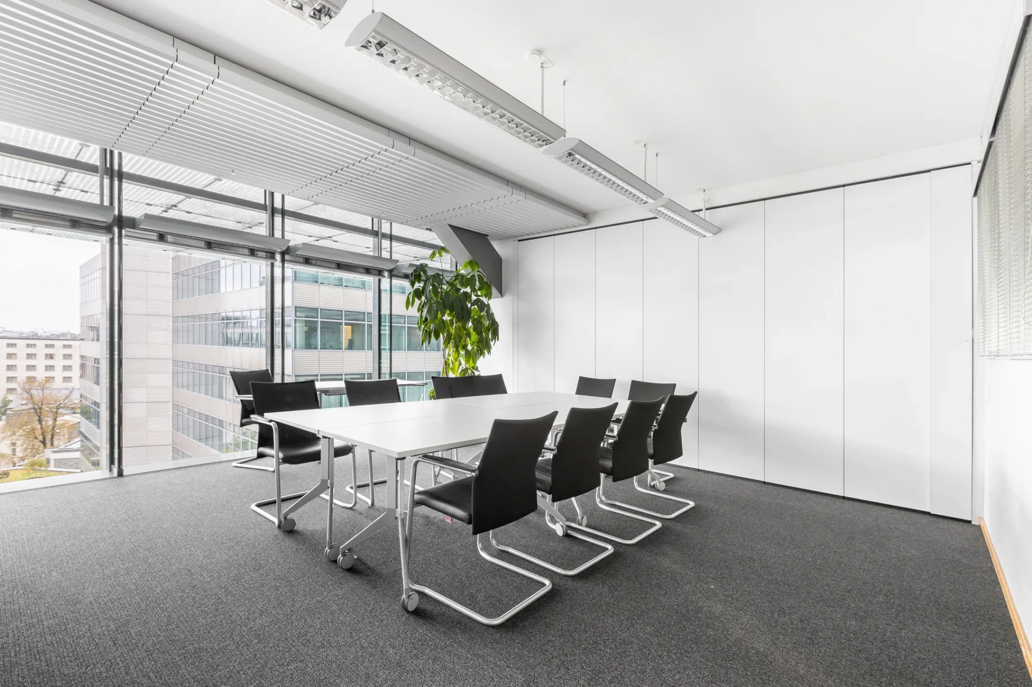 Moderne vergaderruimte met een witte tafel, zwarte stoelen en grote ramen met uitzicht op kantoorgebouwen.