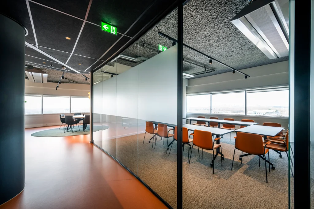 Een moderne vergaderruimte met glazen wanden, oranje stoelen en een grote tafel, verlicht door natuurlijk licht van grote ramen.
