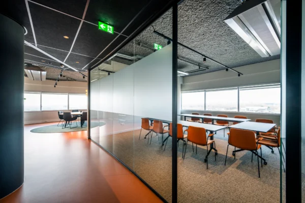 Een moderne vergaderruimte met glazen wanden, oranje stoelen en een grote tafel, verlicht door natuurlijk licht van grote ramen.