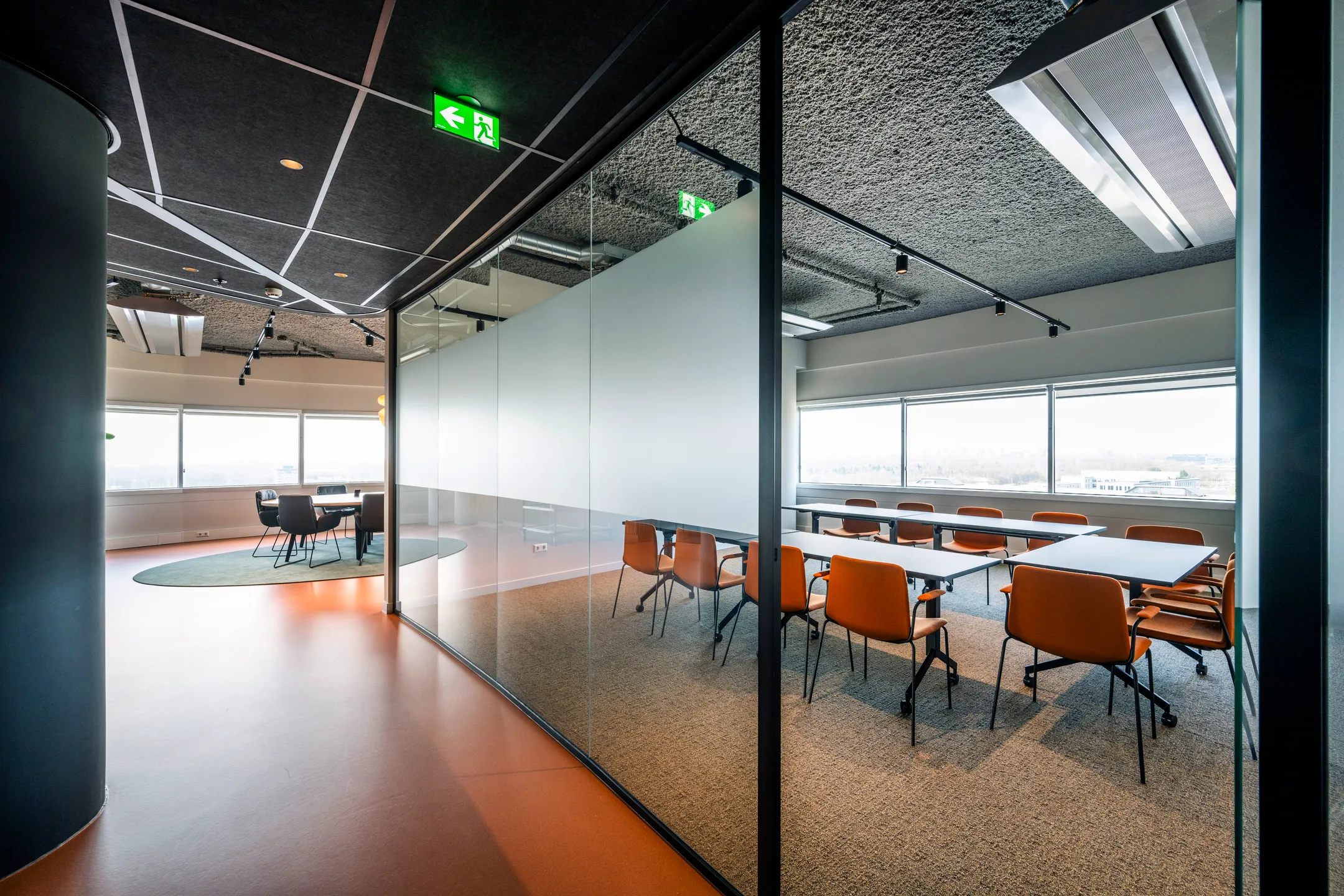 Een moderne vergaderruimte met glazen wanden, oranje stoelen en een grote tafel, verlicht door natuurlijk licht van grote ramen.
