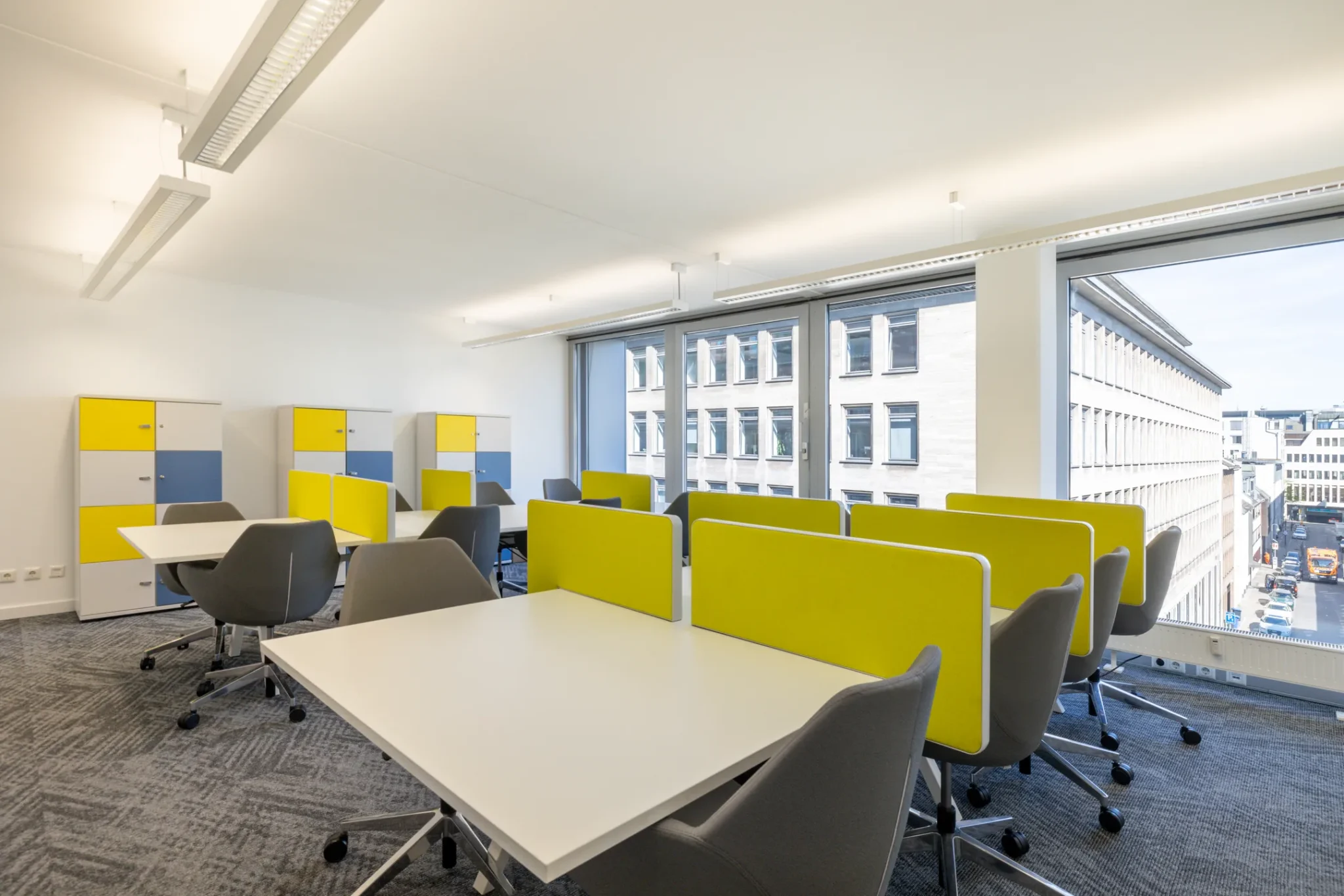 Moderne kantoorruimte met bureaus, grijze stoelen en gele scheidingspanelen, gelegen in een lichte ruimte met grote ramen en uitzicht op de stad.