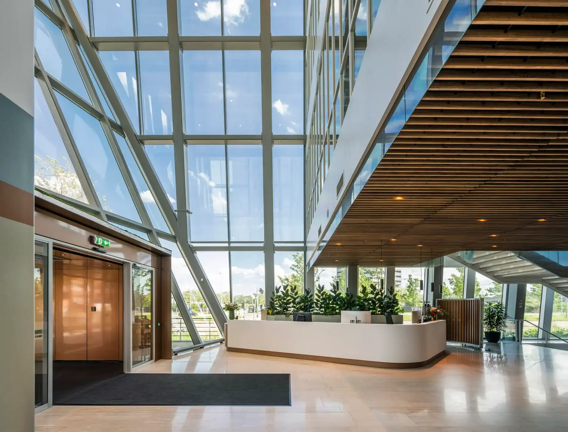 Moderne entreehal met glazen gevel, houten plafond en een receptiebalie met planten.