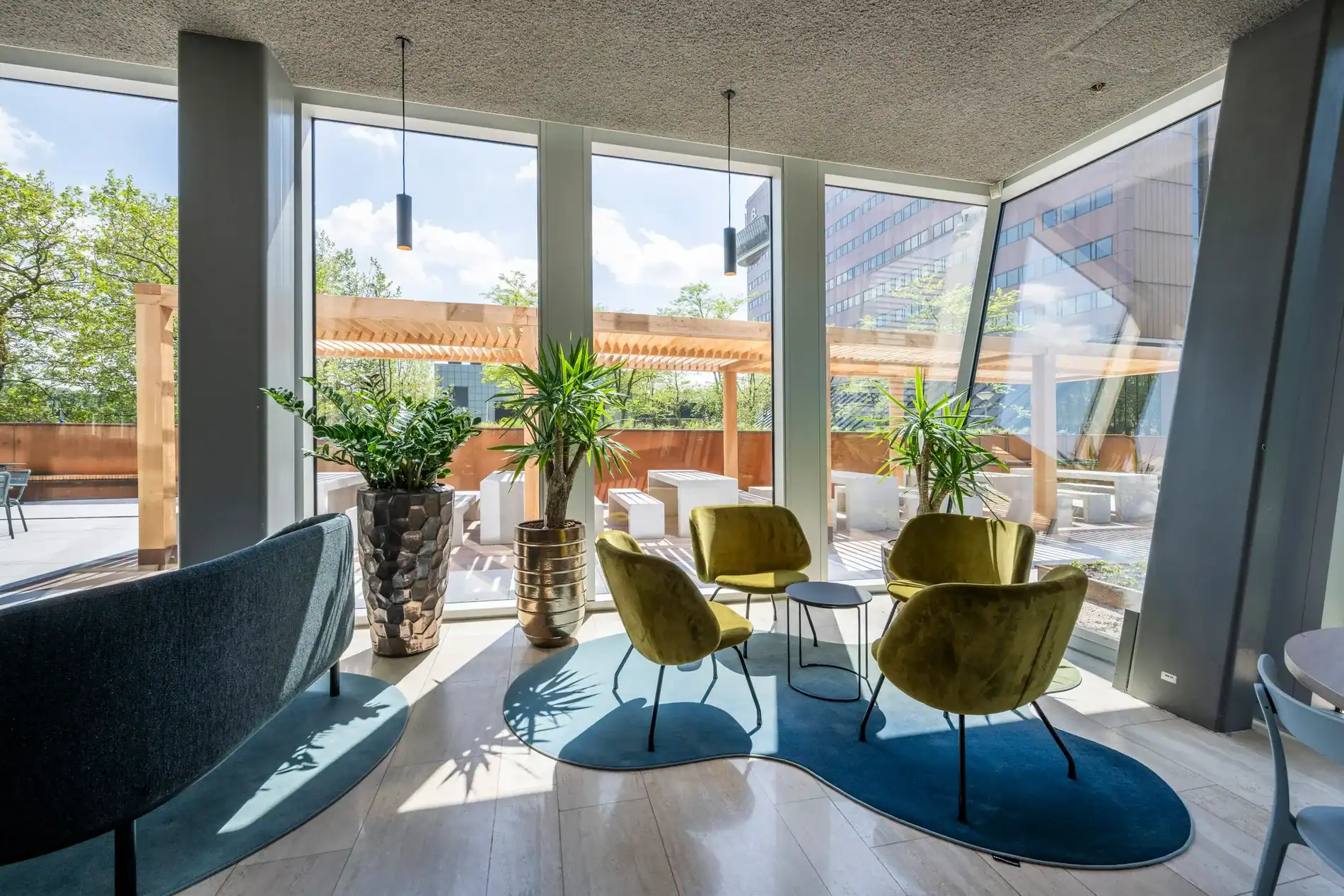 Moderne zithoek met groene stoelen, planten en uitzicht op een zonnig terras met houten pergola.