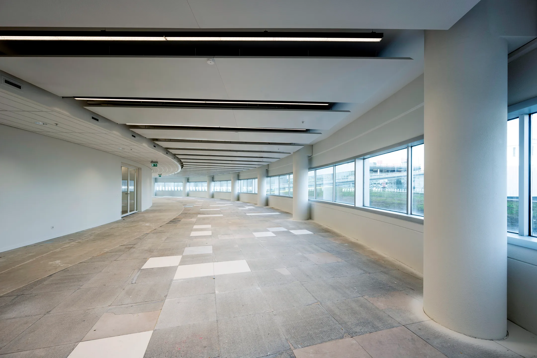 Een lege, licht gebogen gang met grote ramen en een betegelde vloer in een modern gebouw.