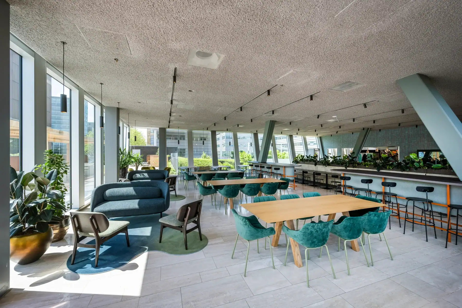 Modern restaurantinterieur met groenfluwelen stoelen, houten tafels en grote ramen met uitzicht op een stedelijke omgeving.