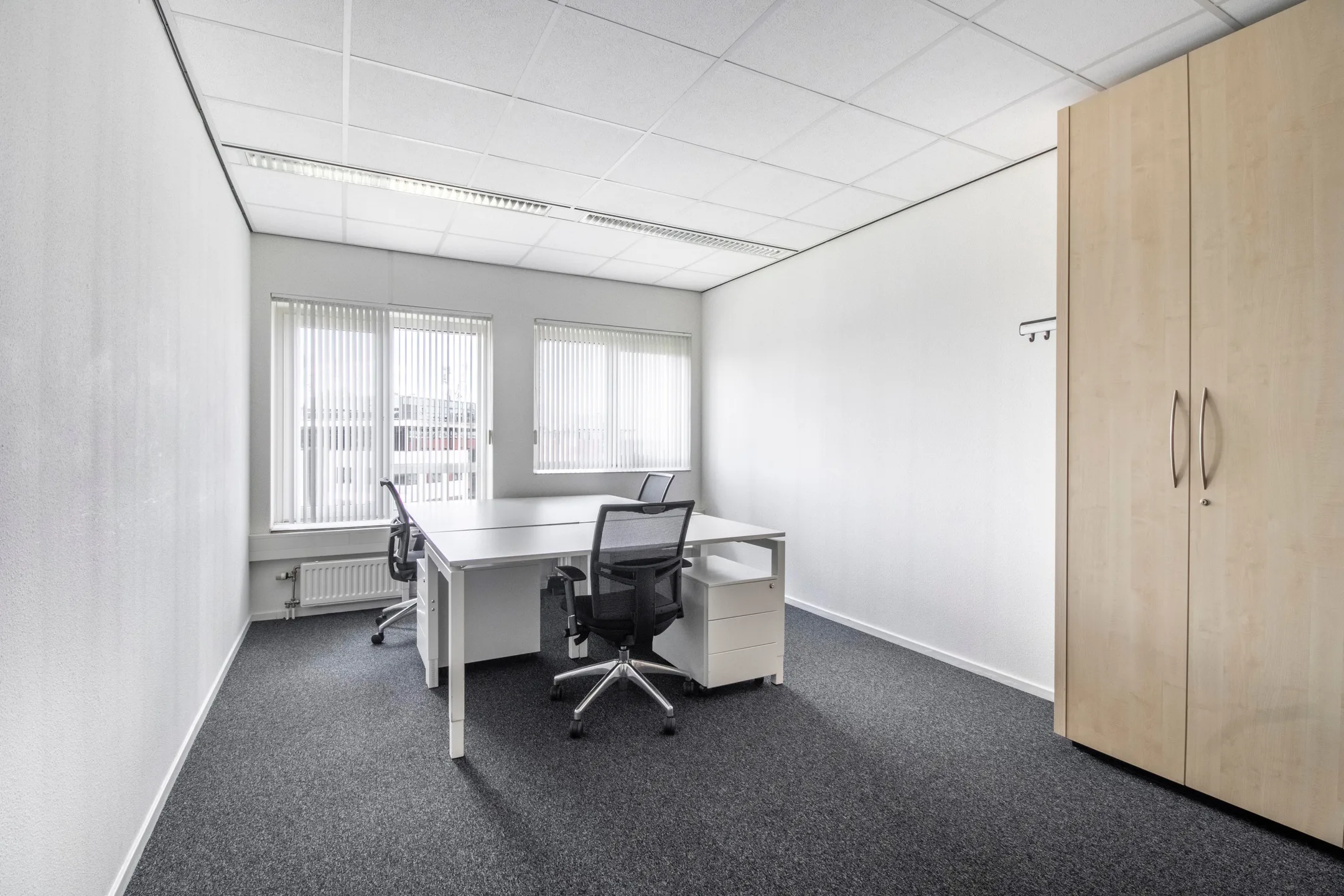 Een lege, moderne kantoorruimte met twee bureaus, bureaustoelen en een kledingkast.