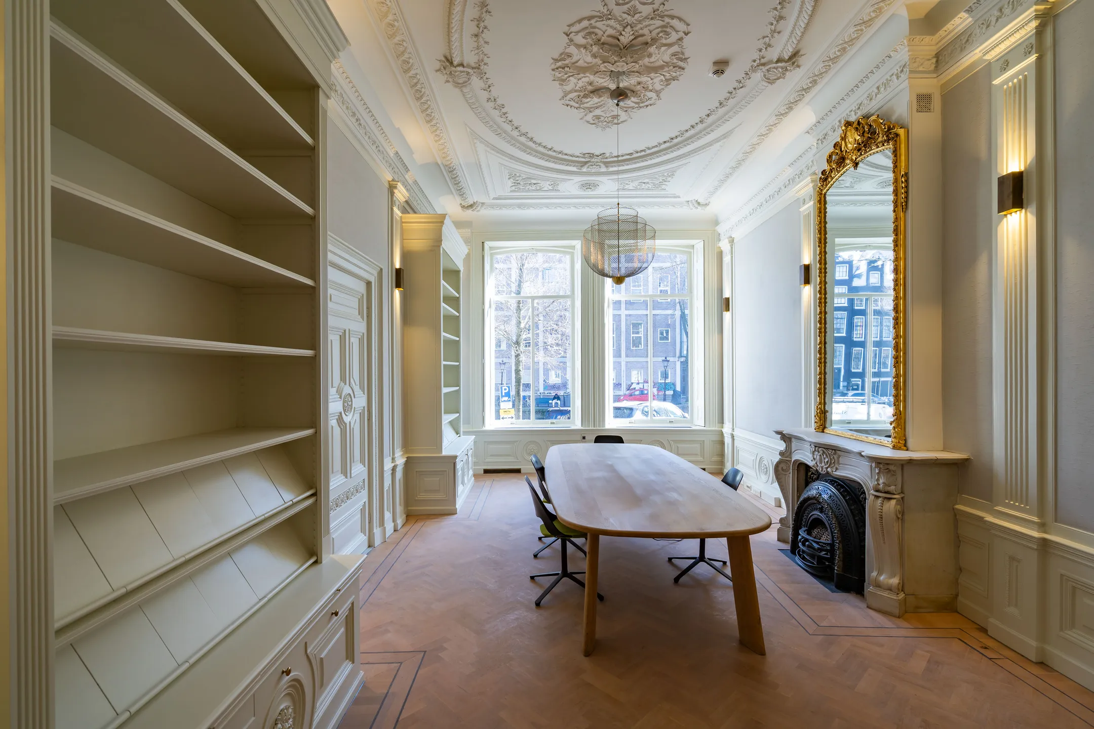 Elegante vergaderruimte met een lange houten tafel, sierlijke plafonddecoraties en een grote gouden spiegel boven de open haard.