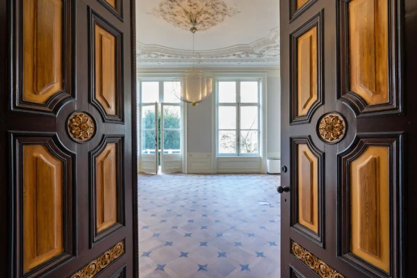 Dubbele deuren openen naar een ruime, sierlijke kamer met grote ramen en een decoratief plafond.