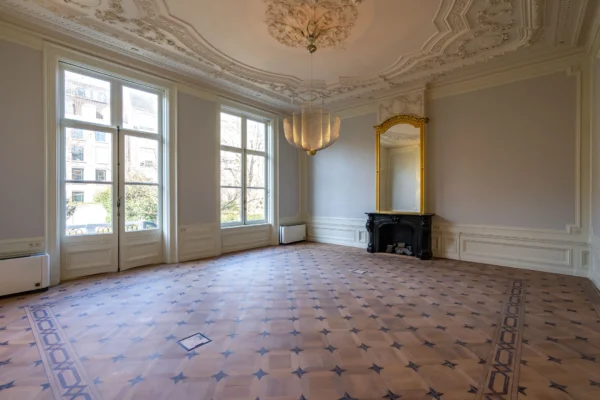 Een ruime, elegante kamer met een sierplafond, grote ramen, een antieke spiegel en een decoratieve houten vloer.