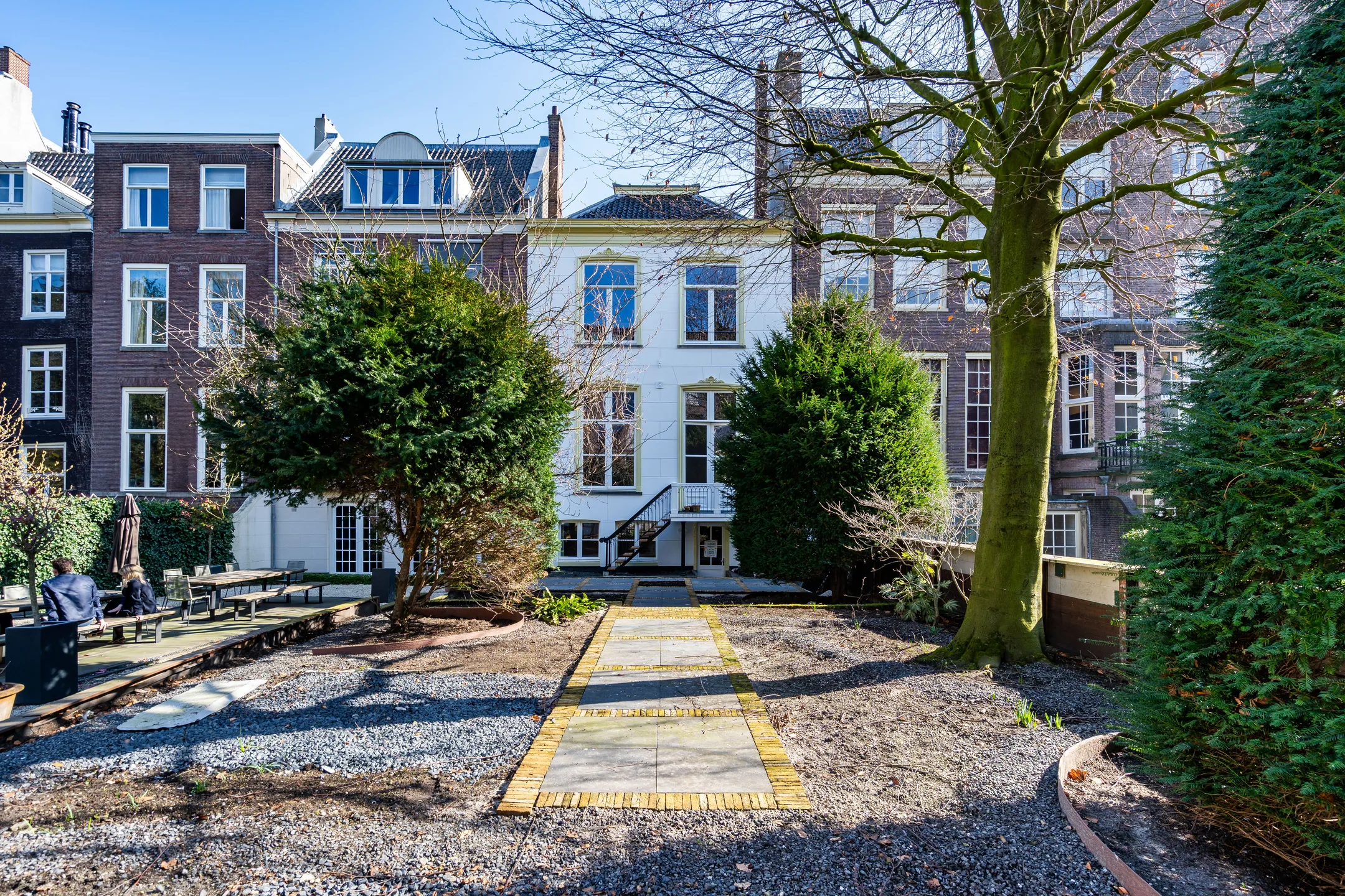 Achtertuin van een klassiek rijtjeshuis met stenen pad, geflankeerd door bomen en struiken.