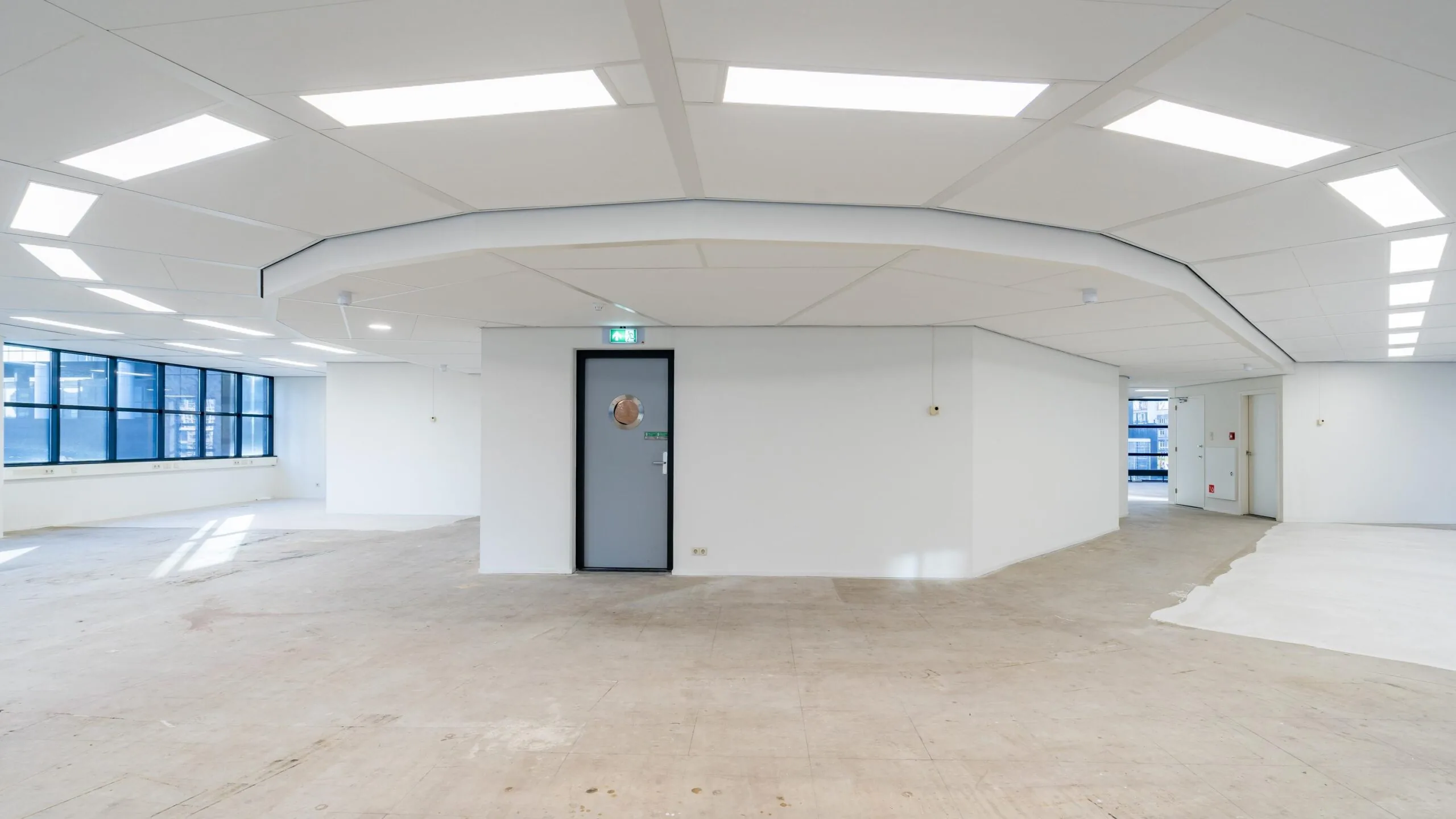 Lege kantoorruimte met witte muren, ramen aan de linkerkant en een grijze deur in het midden van de muur.