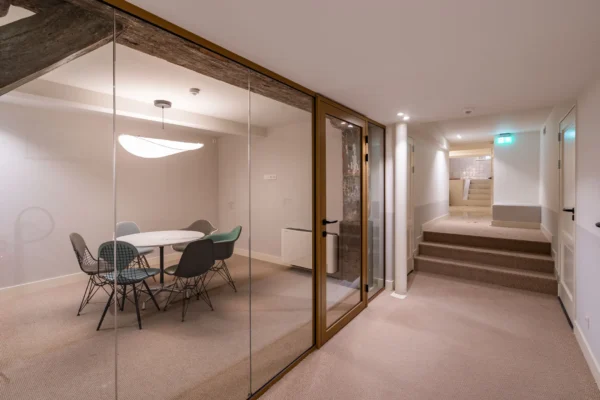 Een moderne vergaderruimte met glazen wand en ronde tafel met stoelen in een verlichte gang.