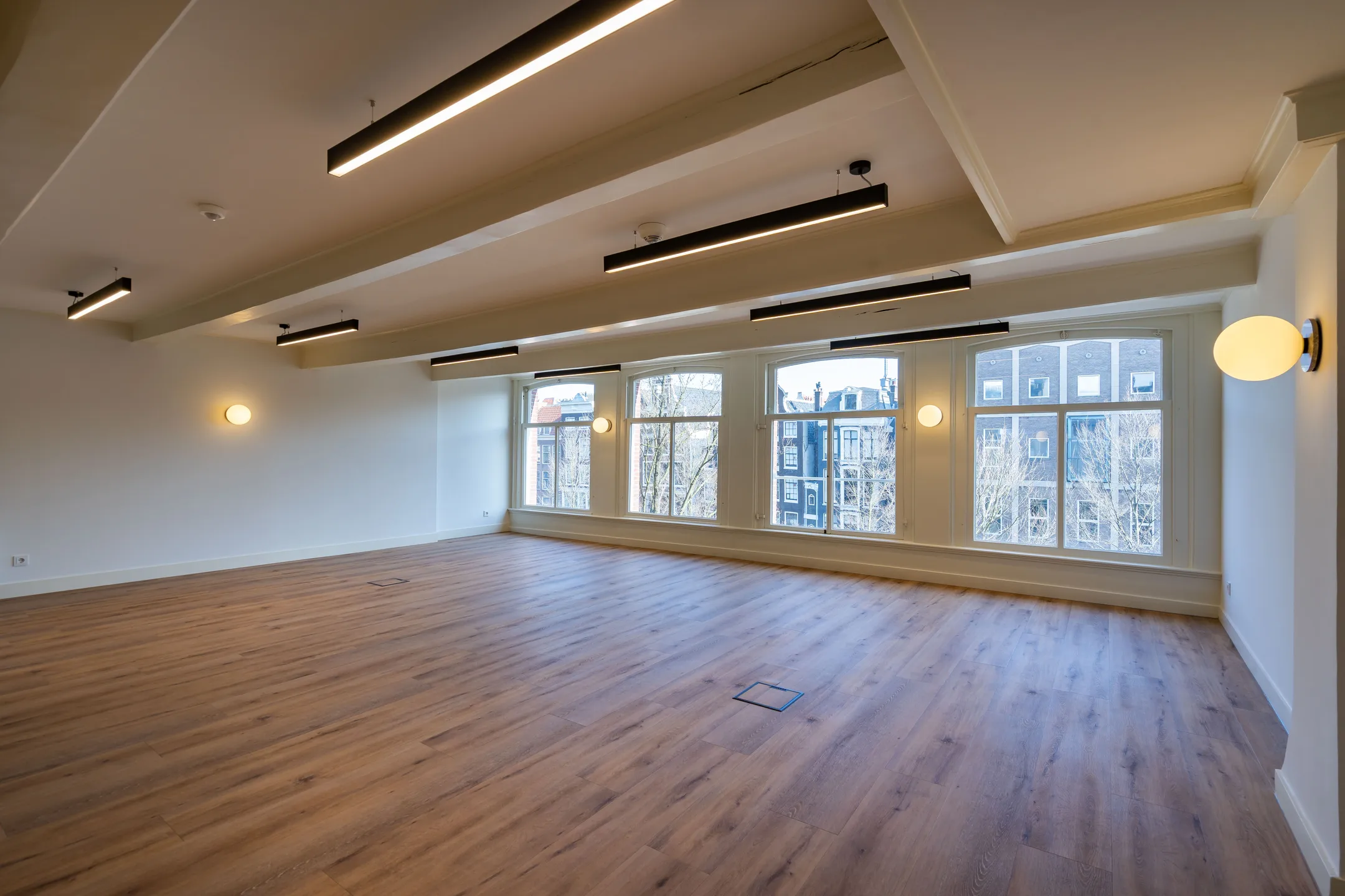 Lege kantoorruimte met houten vloer en grote ramen die uitkijken op gebouwen buiten.