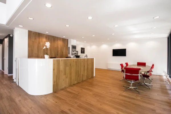 Een moderne receptieruimte met een houten vloer, een witte balie met een bloem en rode stoelen rond een tafel.