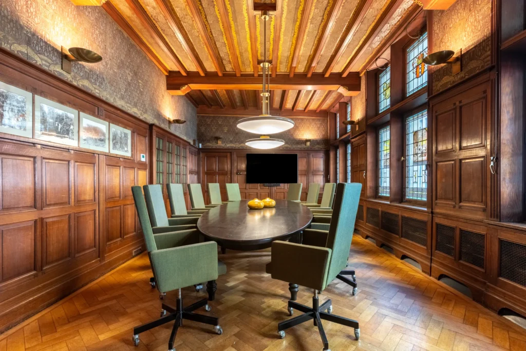Een elegante vergaderruimte met een ovale houten tafel, omgeven door groene stoelen en decoratieve houten panelen.