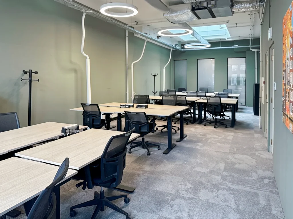 Een moderne vergaderruimte met meerdere bureaus en zwarte stoelen onder ronde plafondlampen.