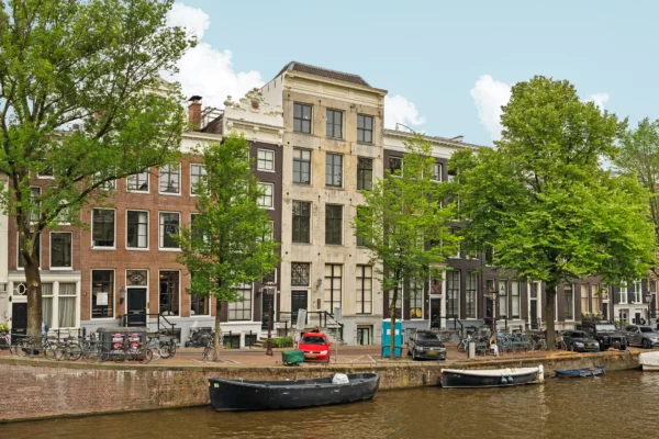Rij historische grachtenpanden aan een Amsterdamse gracht, omgeven door bomen en fietsen.