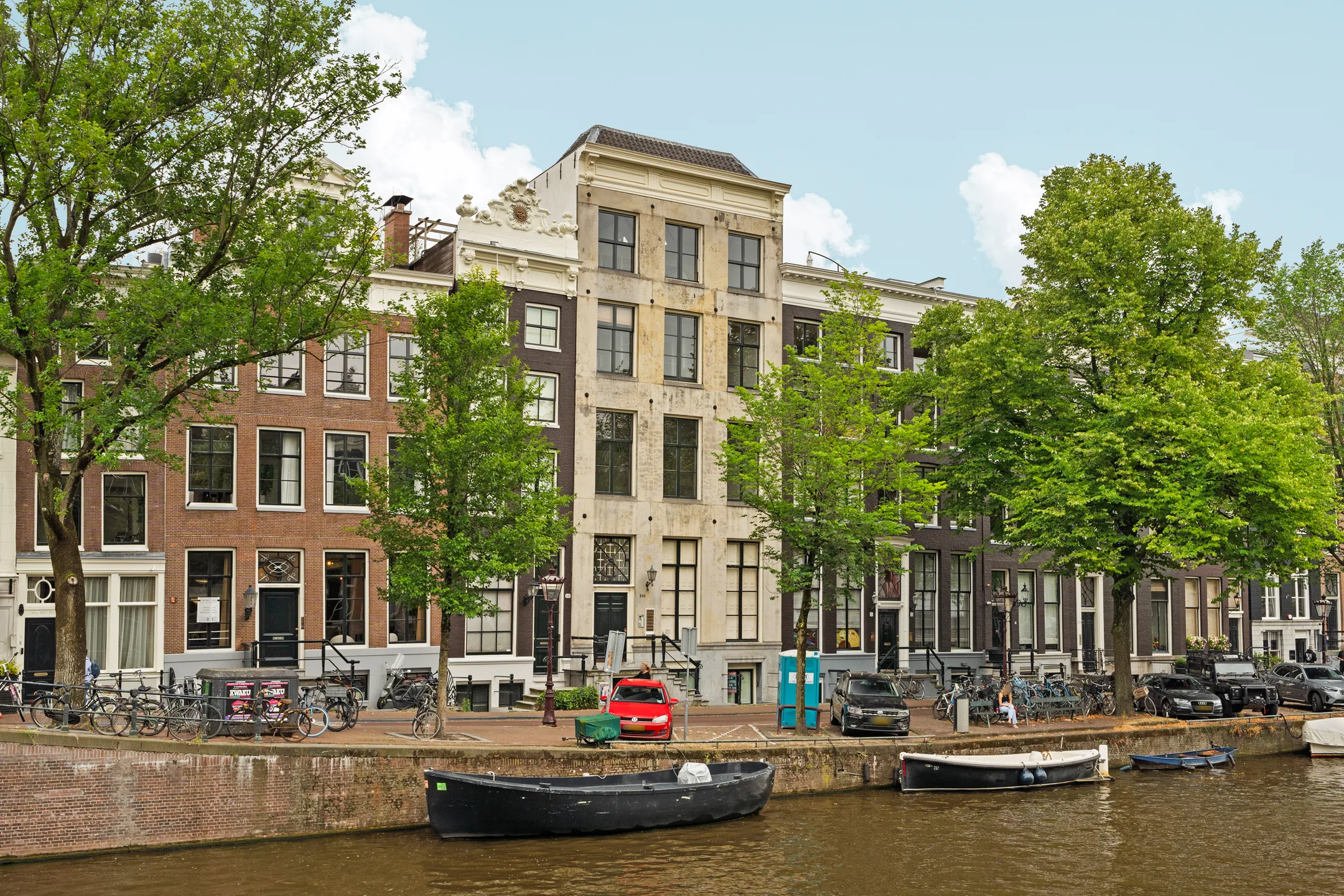Rij historische grachtenpanden aan een Amsterdamse gracht, omgeven door bomen en fietsen.