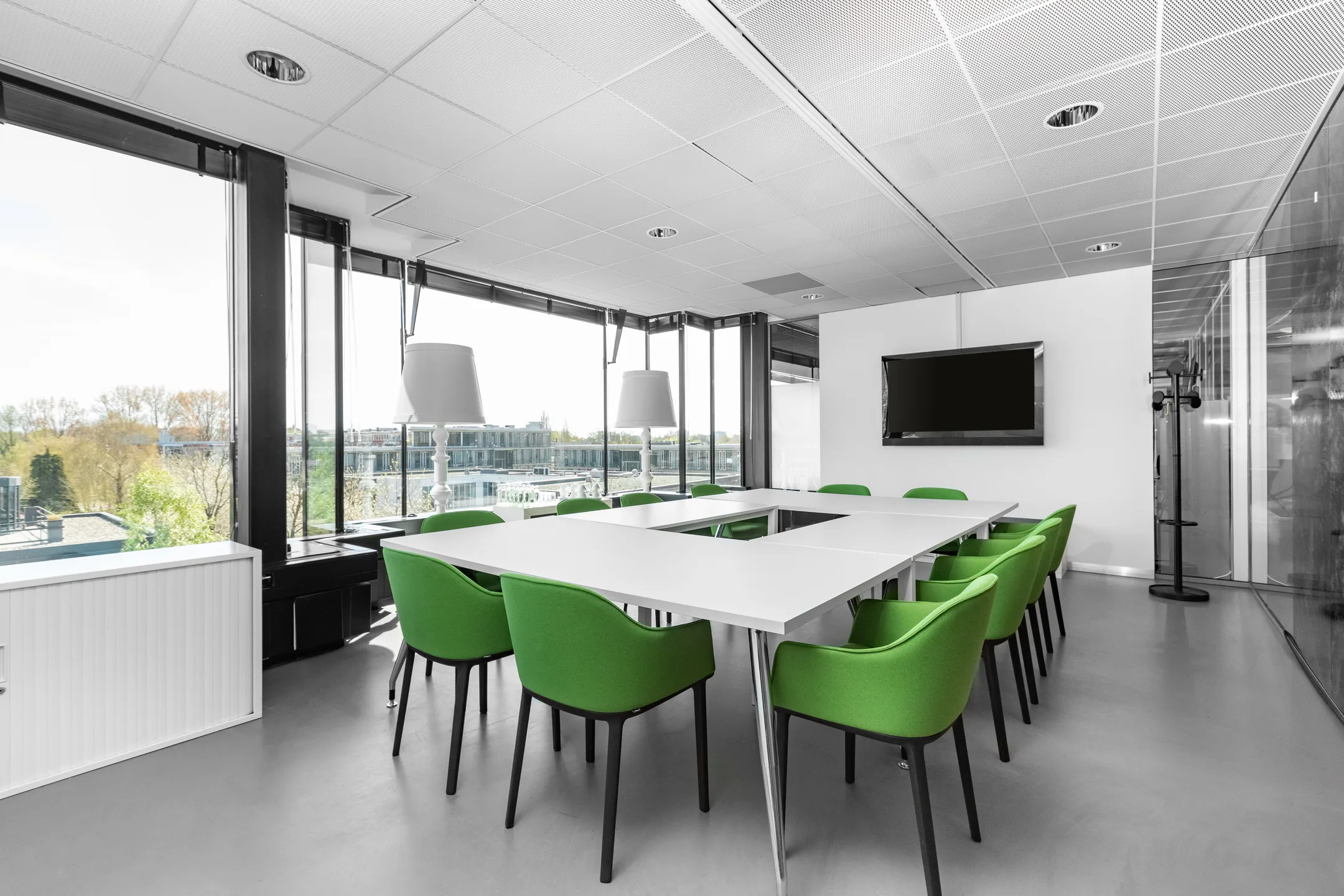 Een moderne vergaderruimte met een tafel omringd door groene stoelen, grote ramen en een flatscreen aan de muur.
