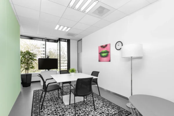 Een modern ingerichte vergaderruimte met een rechthoekige tafel, stoelen, een zwarte tv, een groene plant, en een kunstwerk van felgroene lippen aan de muur.