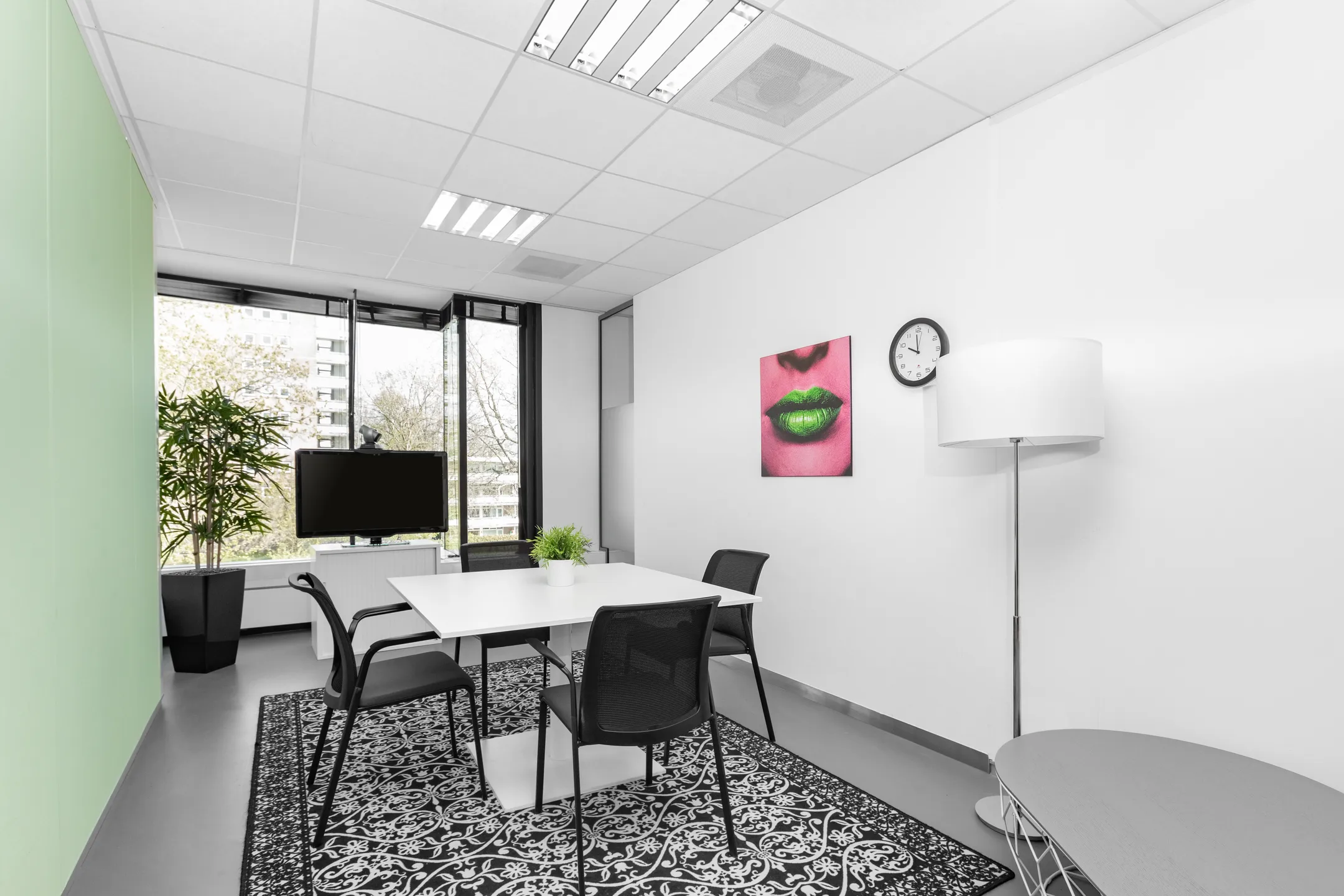 Een modern ingerichte vergaderruimte met een rechthoekige tafel, stoelen, een zwarte tv, een groene plant, en een kunstwerk van felgroene lippen aan de muur.