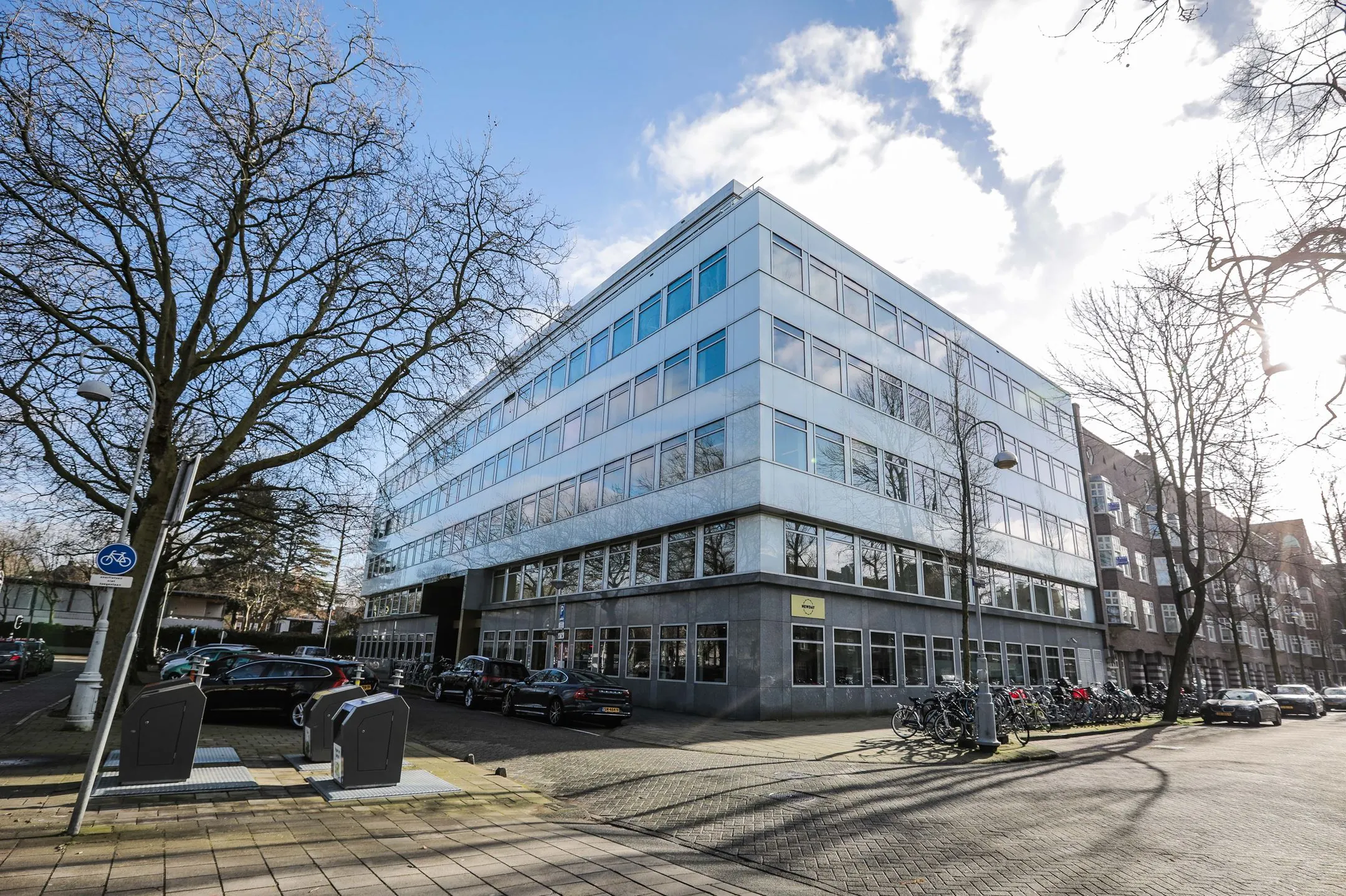 Modern kantoorgebouw op een straathoek met geparkeerde auto's en fietsen, onder een heldere blauwe lucht.