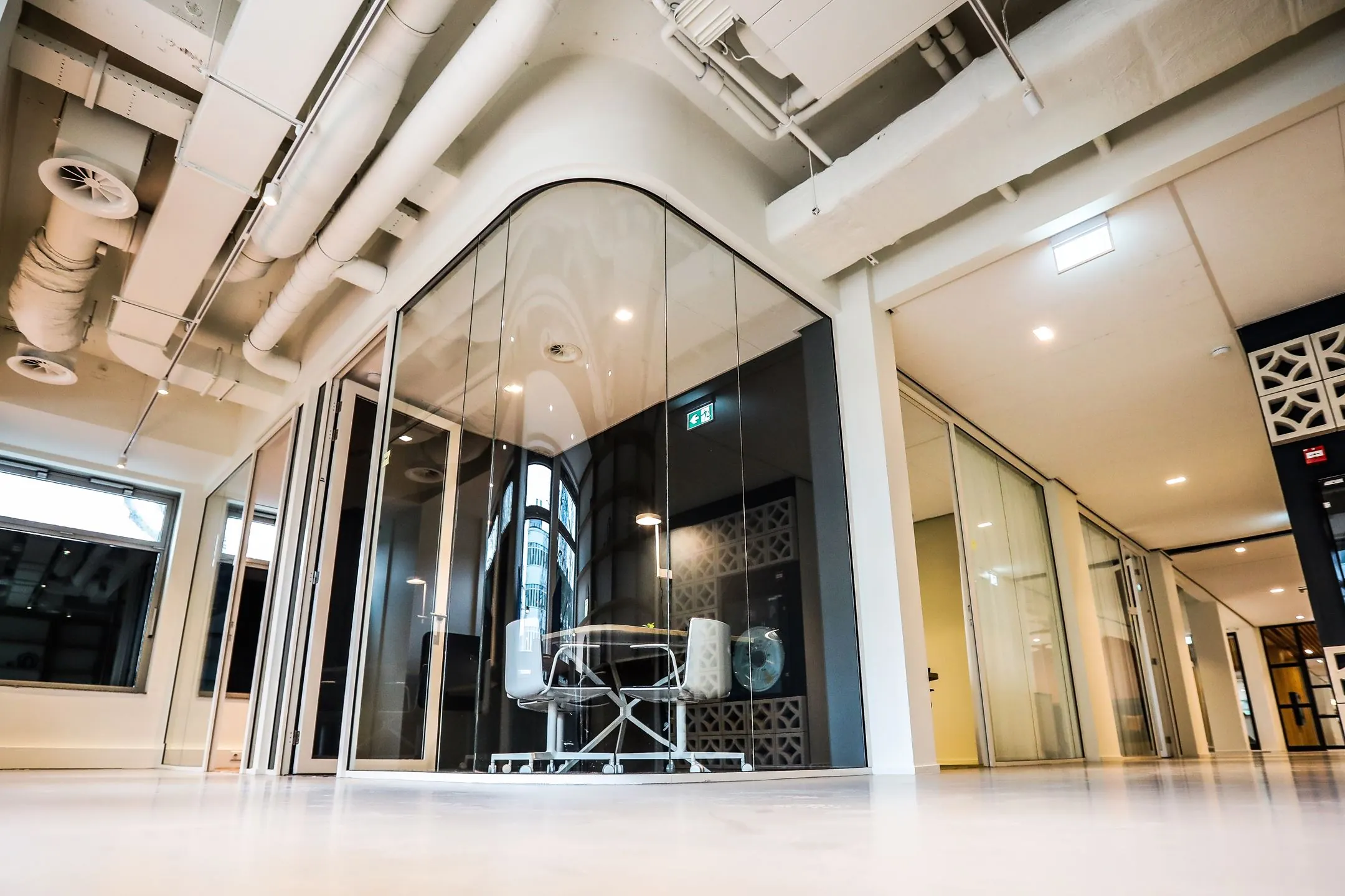 Modern kantoorinterieur met glazen vergaderruimte, witte buizen aan het plafond en strakke afwerkingen.