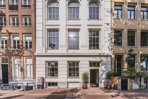Een historische gevel van een pand tussen twee andere gebouwen met schaduwen van bomen op de gevels.
