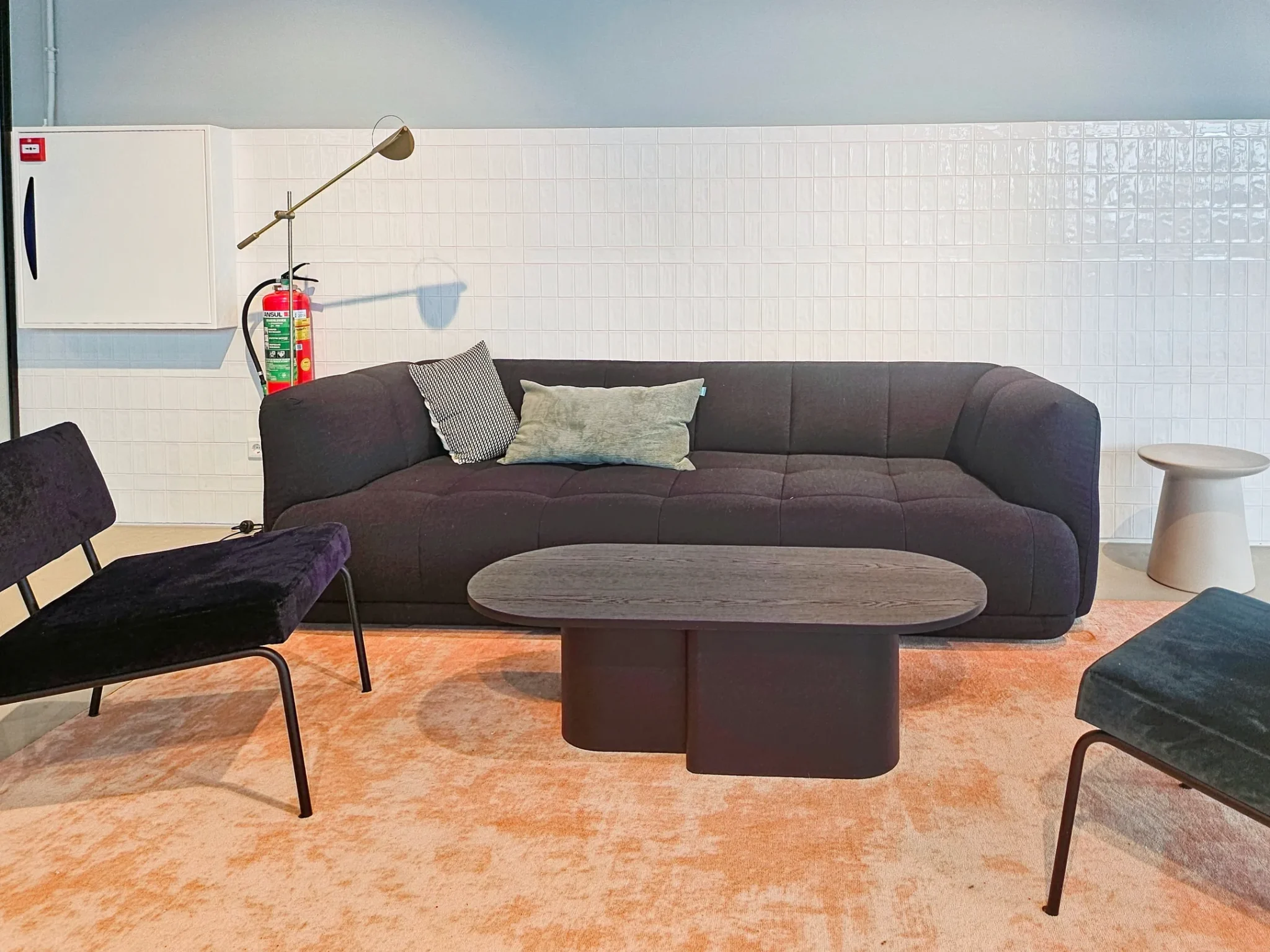 Moderne zithoek met een donkerpaarse bank, zwarte stoelen, een ovale salontafel en een lichtoranje vloerkleed tegen een witte betegelde wand.