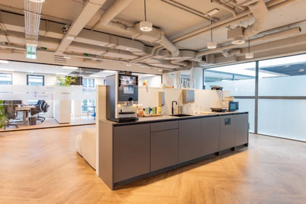 Moderne kantoorruimte met een kitchenette voorzien van een koffiezetapparaat, glazen, en een magnetron, naast een werkruimte met glazen wanden.