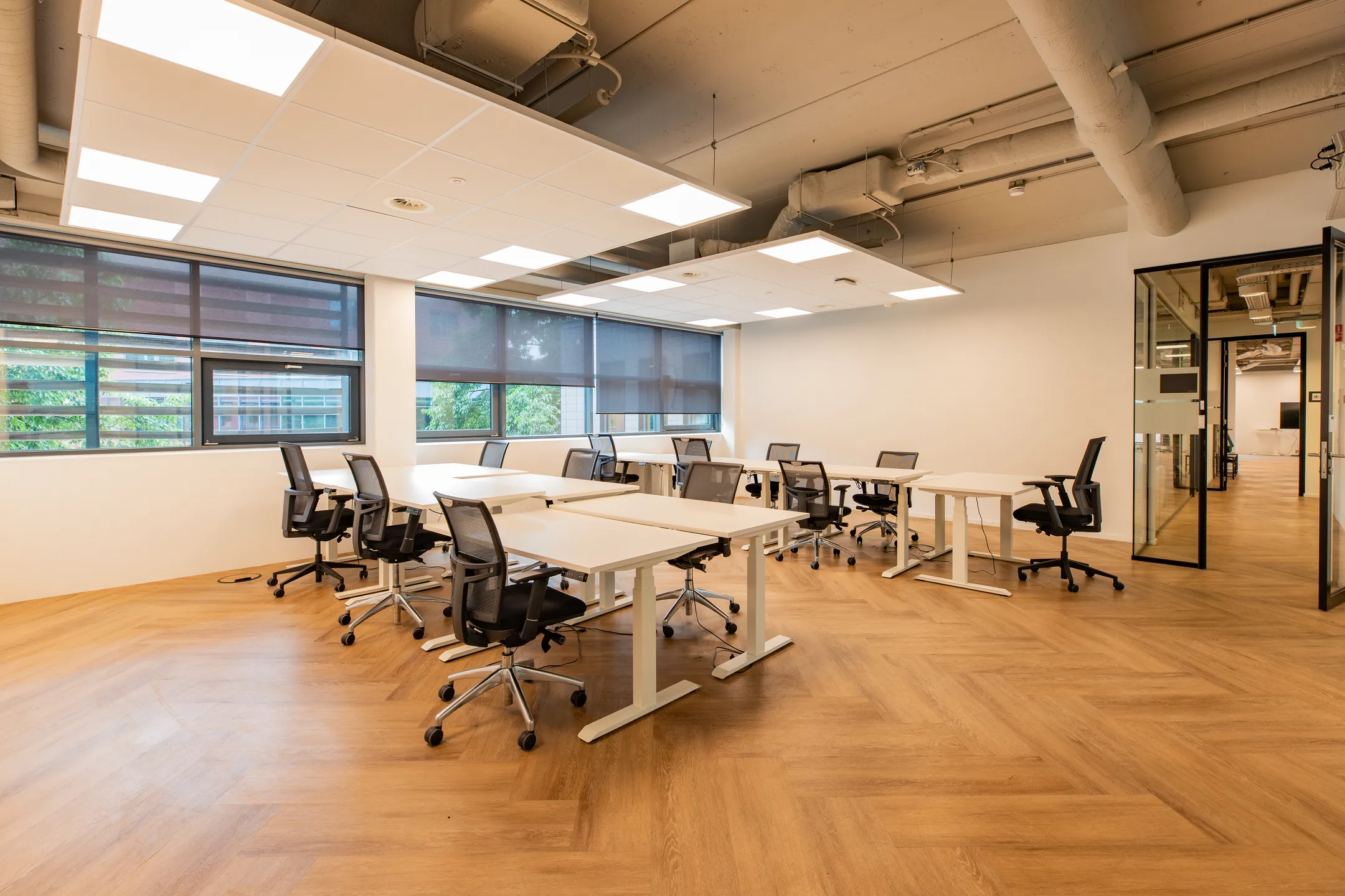 Moderne kantoorruimte met bureaus en stoelen, omgeven door grote ramen en houten vloeren.