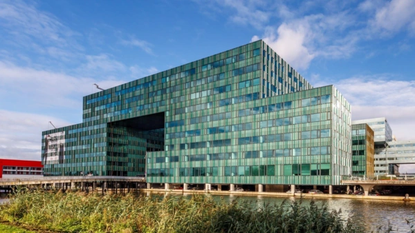 Modern kantoorgebouw aan de Herikerbergweg in Amsterdam, met groene glazen gevel en overhangende constructie boven het water.