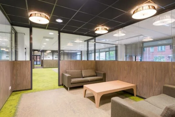 Moderne kantoorruimte met glazen wanden, groene vloerbedekking en een zithoek met bruine banken en houten tafel.