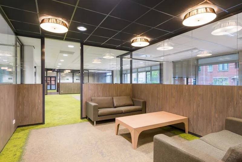 Moderne kantoorruimte met glazen wanden, groene vloerbedekking en een zithoek met bruine banken en houten tafel.