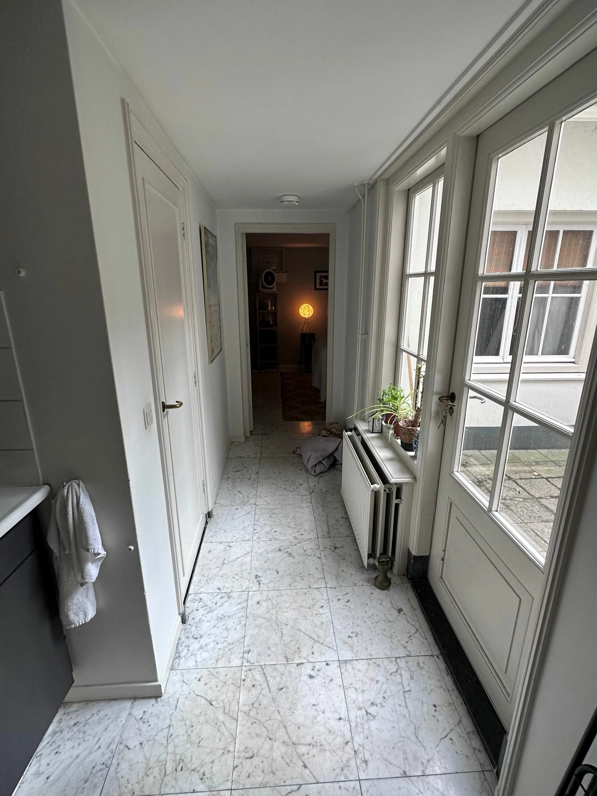 Een smalle, lichte gang met marmeren vloer, een open deur naar een kamer met een brandende lamp en een radiator onder het raam.