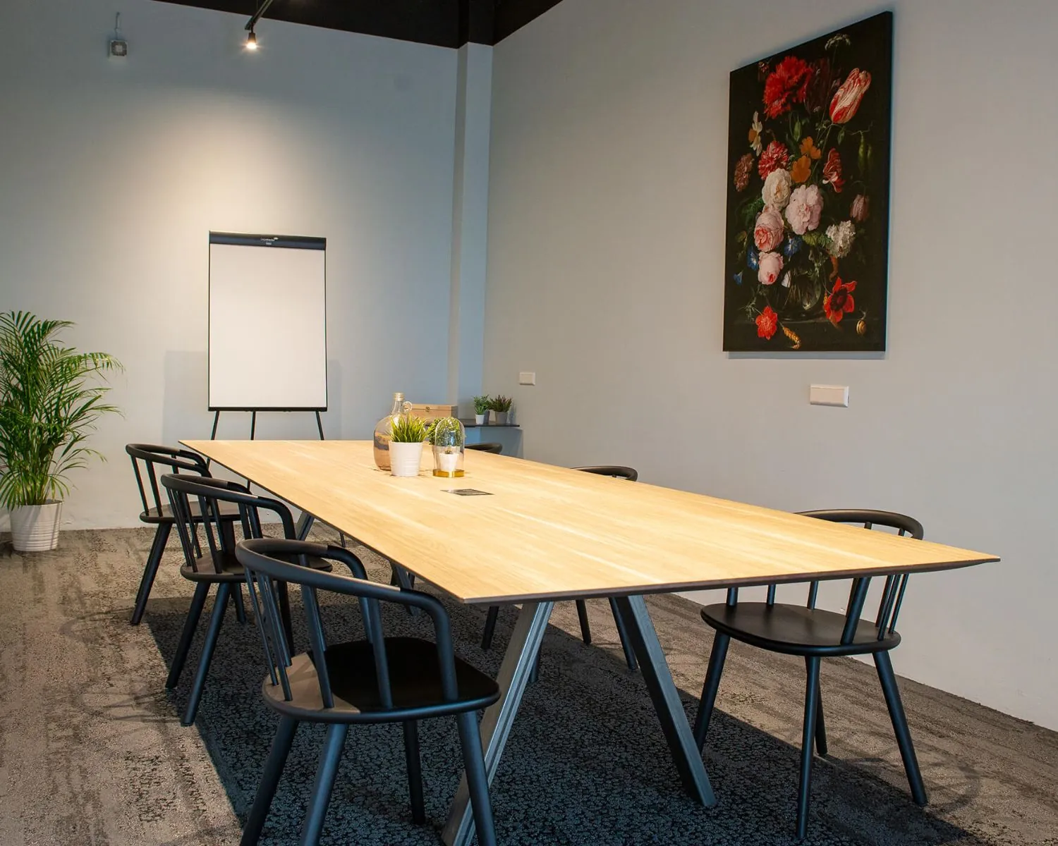 Vergaderruimte met een houten tafel, zwarte stoelen, flipover en een schilderij van bloemen aan de muur.